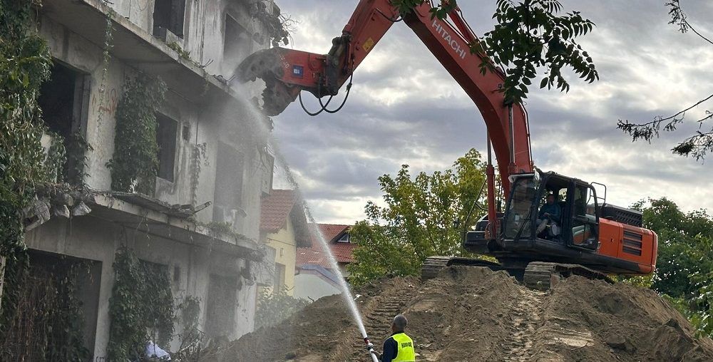 Excavadora Hitachi con pinza demoledora derribando un edificio de hormigón armado de varias plantas, con riego de agua para controlar el polvo