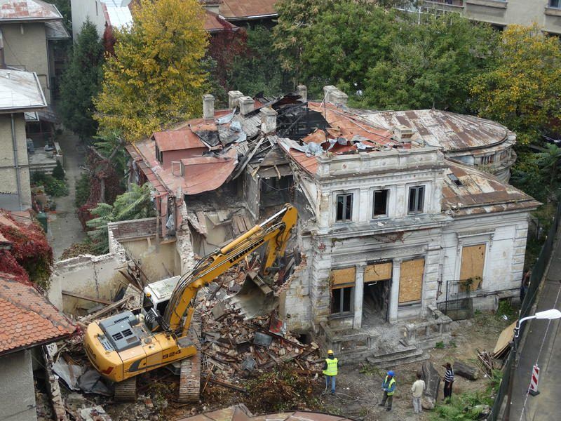 Vista aérea: excavadora Liebherr demoliendo una antigua villa en un entorno urbano denso